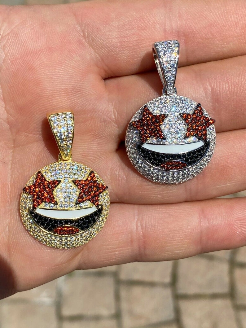 Puede incluir: Dos colgantes de oro y plata con un dise&ntilde;o de cara sonriente. La cara sonriente tiene ojos en forma de estrella roja y una boca negra. Los colgantes est&aacute;n cubiertos de piedras de circonita c&uacute;bica transparentes y negras.