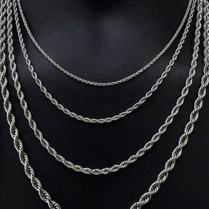 May include: A selection of silver rope chain necklaces in different thicknesses, presented on a black mannequin. The necklaces are layered, highlighting the various sizes and textures of the chains. The chains are made from a shiny, metallic material.