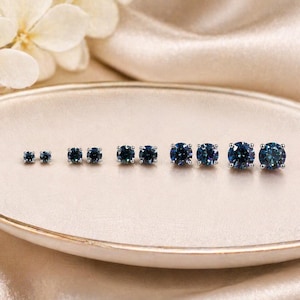 May include: A collection of blue gemstone stud earrings of varying sizes, displayed on a light-colored, oval-shaped plate. The earrings are arranged in a row, showcasing the different sizes and the sparkle of the gemstones. The background is a soft, neutral color.