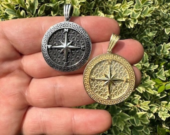 May include: Two round pendant necklaces with compass rose designs. One is silver-toned, the other gold-toned. Both have a decorative border and a textured background. The pendants are held in front of a green leafy background.