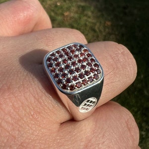May include: A silver ring with a square face, densely set with small, round, deep red gemstones. The ring has a polished finish and a geometric design on the side.