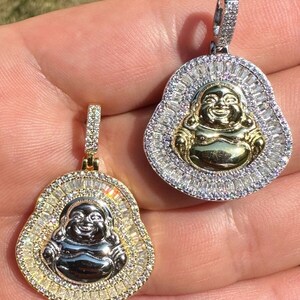 May include: Two gold and silver Buddha pendants. Each pendant features a smiling Buddha figure at the centre, surrounded by a halo of sparkling stones. The pendants are held up by a hand, showcasing their intricate design and craftsmanship.
