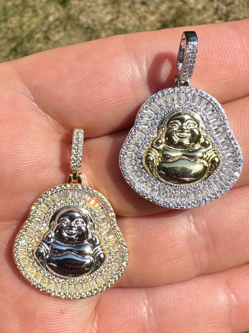 May include: Two gold and silver Buddha pendants. Each pendant features a smiling Buddha figure at the centre, surrounded by a halo of sparkling stones. The pendants are held up by a hand, showcasing their intricate design and craftsmanship.