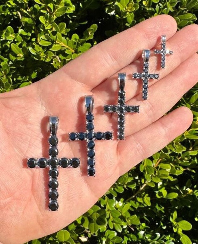 May include: Five silver cross pendants with black cubic zirconia stones. The crosses are of different sizes and are arranged in a row on a hand.