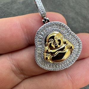 May include: A gold-coloured Buddha pendant with a halo of small, clear stones. The pendant has a silver-coloured bail and a gold-coloured Buddha face. The pendant is likely made of metal and is designed to be worn as jewellery.