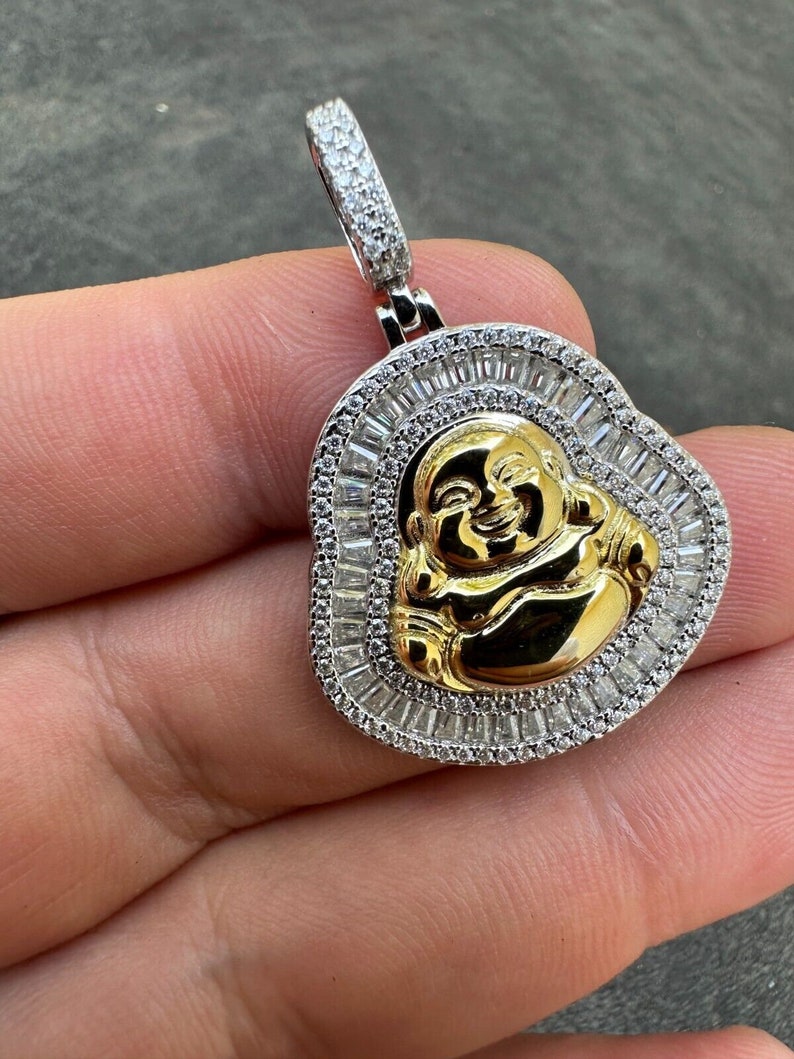 May include: A gold-coloured Buddha pendant with a halo of small, clear stones. The pendant has a silver-coloured bail and a gold-coloured Buddha face. The pendant is likely made of metal and is designed to be worn as jewellery.