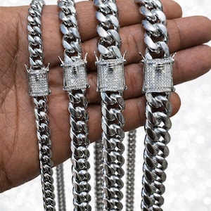 Puede incluir: Se muestran varias cadenas de eslabones cubanos de color plateado de diferentes anchos. Cada cadena presenta un cierre cuadrado adornado con pequeñas piedras transparentes. Las cadenas se sostienen contra un fondo borroso y brillante.