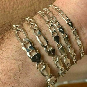 May include: Five silver chain bracelets with a Figaro link pattern. The bracelets are stacked on a person's wrist. The bracelets are marked with "925 Italy" and "925 LUX".