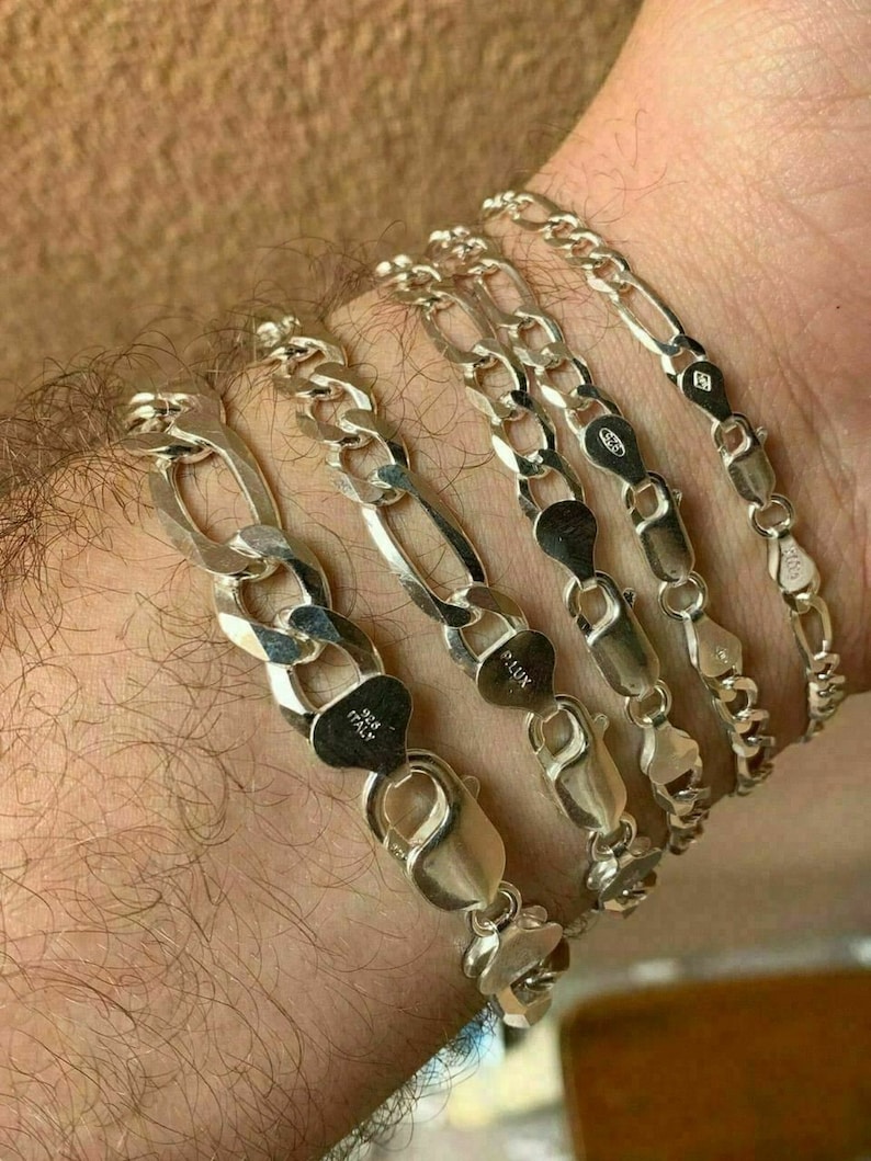 May include: Five silver chain bracelets with a Figaro link pattern. The bracelets are stacked on a person's wrist. The bracelets are marked with "925 Italy" and "925 LUX".