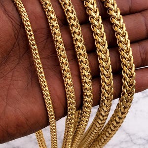 May include: A collection of gold-colored chain necklaces with varying thicknesses. The chains are displayed against a dark skin tone, showcasing their intricate woven designs. The necklaces are a bright, shiny gold.