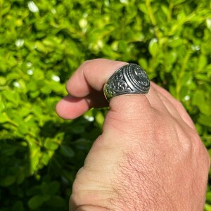 Puede incluir: Un anillo de plata con un dise&ntilde;o adornado y una pieza central circular elevada. El anillo est&aacute; en un dedo.