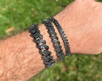 Puede incluir: Tres pulseras negras con diamantes se muestran en la muñeca de una persona. Las pulseras tienen un diseño de eslabones cubanos. Las pulseras son de metal oscuro y están sobre un fondo verde y herboso.