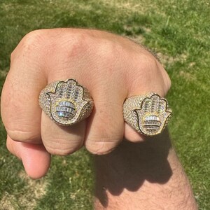 Puede incluir: Dos anillos de oro con un dise&ntilde;o de mano de hamsa engastados con diamantes baguette y redondos transparentes.