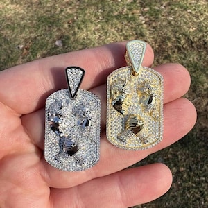 Puede incluir: Dos colgantes de tono dorado con un diseño de cabeza de toro, ambos cubiertos de piedras de circonita cúbica transparentes. Los colgantes tienen la forma de una placa de identificación.