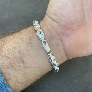 Puede incluir: Un brazalete de cadena de plata con un dise&ntilde;o trenzado. El brazalete est&aacute; en la mu&ntilde;eca de una persona.