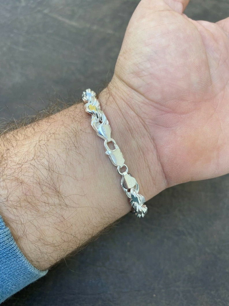 Puede incluir: Un brazalete de cadena de plata con un dise&ntilde;o trenzado. El brazalete est&aacute; en la mu&ntilde;eca de una persona.