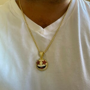 Puede incluir: Un collar de cadena de oro con un colgante de oro que presenta una cara sonriente con ojos en forma de estrella roja y dientes blancos. El colgante est&aacute; cubierto de peque&ntilde;as piedras preciosas transparentes y rojas.
