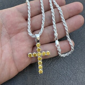 May include: A silver rope chain necklace with a silver cross pendant. The cross is encrusted with yellow cubic zirconia stones.
