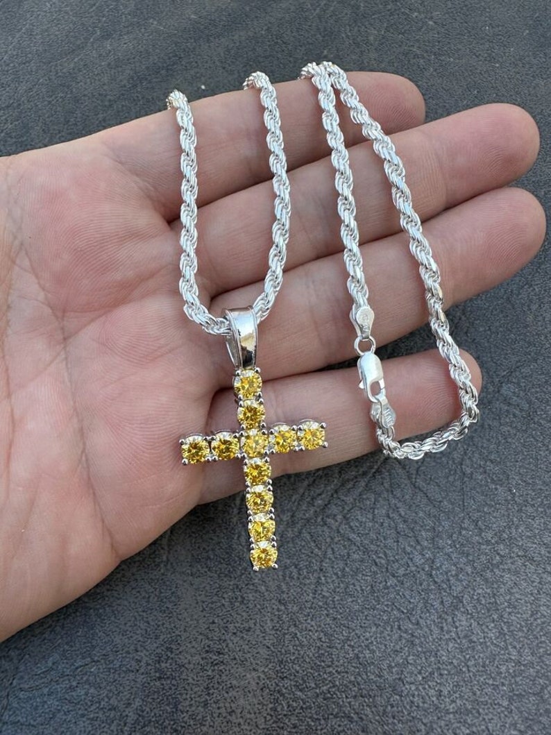 May include: A silver rope chain necklace with a silver cross pendant. The cross is encrusted with yellow cubic zirconia stones.