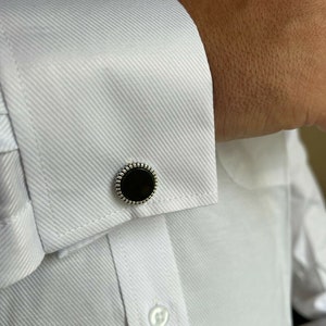 Op de afbeelding: Close-up van een wit overhemd met een textuurpatroon en een zwarte en zilveren manchetknop. De manchetknop heeft een ronde, zwarte kern met een decoratieve zilveren rand. Het overhemd is geknoopt en heeft een klassieke kraag.