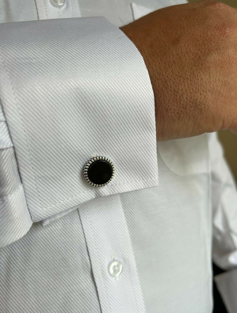 Op de afbeelding: Close-up van een wit overhemd met een textuurpatroon en een zwarte en zilveren manchetknop. De manchetknop heeft een ronde, zwarte kern met een decoratieve zilveren rand. Het overhemd is geknoopt en heeft een klassieke kraag.