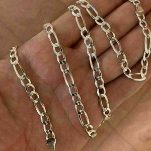 May include: A silver chain necklace with a figaro link pattern. The chain is laid out on a hand, showing the length and detail of the links.
