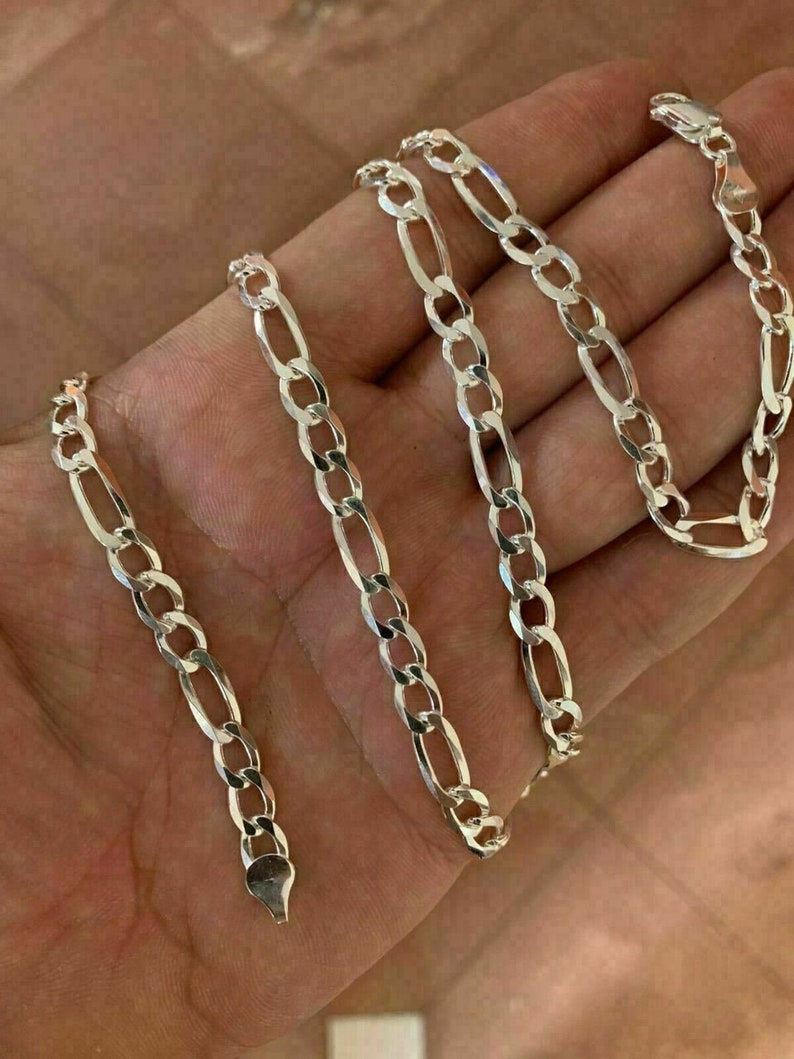 May include: A silver chain necklace with a figaro link pattern. The chain is laid out on a hand, showing the length and detail of the links.