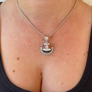 Puede incluir: Un collar de cadena de plata con un gran colgante redondo. El colgante est&aacute; cubierto de peque&ntilde;as piedras preciosas blancas y rojas, formando un dise&ntilde;o de cara sonriente.