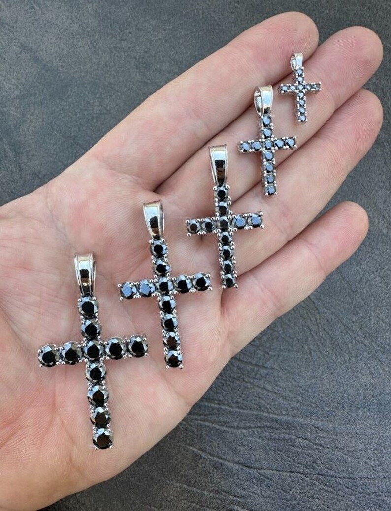 May include: Five silver cross pendants with black diamond accents. The crosses are of varying sizes, with the largest cross at the bottom of the image and the smallest cross at the top.