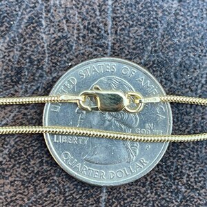 Puede incluir: Un collar de cadena de oro con un cierre de langosta, colocado sobre una moneda de 25 centavos de d&oacute;lar estadounidense. La moneda dice "United States of America" y "Liberty Quarter Dollar".