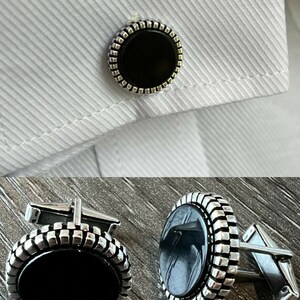 Op de afbeelding: Zilveren manchetknopen met een zwart ovale middenstuk en een getextureerde rand. De manchetknopen worden getoond op een wit overhemd. Klassiek ontwerp geschikt voor formele gelegenheden.