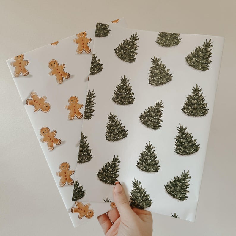 May include: Two sheets of Christmas wrapping paper. One has gingerbread men, the other has green Christmas trees. Both have a white background. The paper is held by a hand.