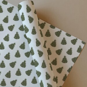 May include: White wrapping paper featuring a repeating pattern of green Christmas trees. The paper is partially rolled, showing the pattern on both sides. The trees have dark green outlines and shading.