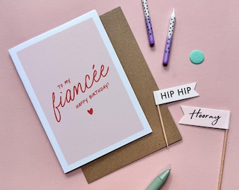 Könnte beinhalten: Eine Geburtstagskarte mit dem Text "To My Fiancee Happy Birthday!" in roter Schrift, ein brauner Umschlag, zwei lila Kerzen mit weißen Punkten und zwei kleine Fähnchen mit "Hip Hip" und "Hooray". Der Hintergrund ist rosa.