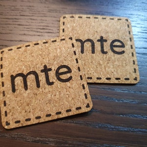 Könnte beinhalten: Zwei braune Korkuntersetzer mit einem schwarzen genähten Rand und dem Wort "mte" in Schwarz gedruckt.