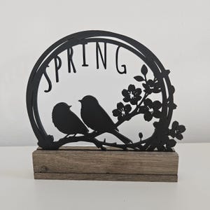 Könnte beinhalten: Schwarze Frühlingsdekoration aus Metall mit zwei Vögeln und einem blühenden Zweig, das Wort "SPRING" ist aus dem Kreis ausgeschnitten, die Dekoration steht auf einem Holzsockel.