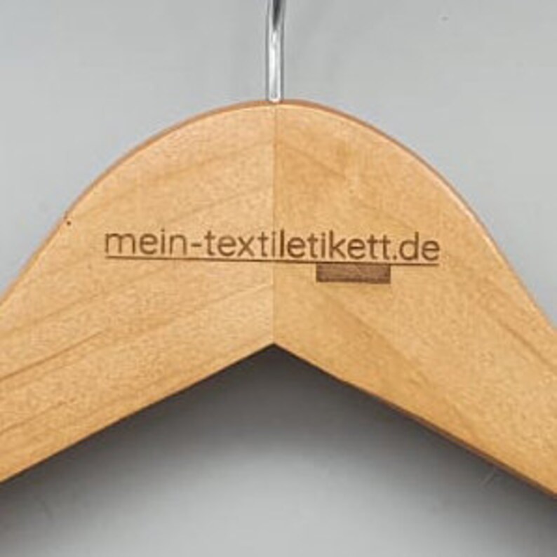 May include: A natural wood hanger with a rounded top and a smooth finish. The hanger has the text "mein-textilticket.de" printed on the front.