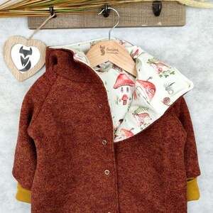 May include: A brown fleece jacket with a removable lining featuring a whimsical mushroom and house print. The lining is white with brown, green, and yellow accents. The jacket has a zipper closure and two pockets.