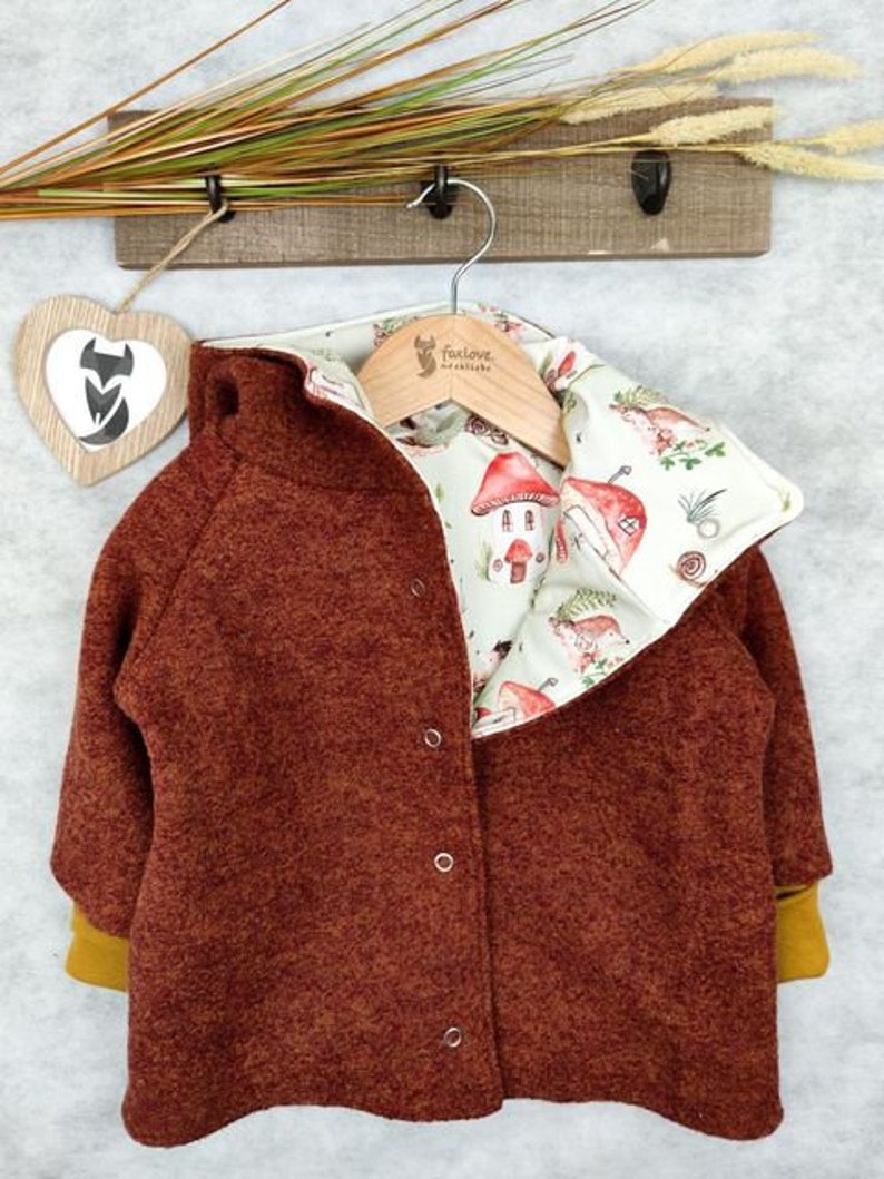 May include: A brown fleece jacket with a removable lining featuring a whimsical mushroom and house print. The lining is white with brown, green, and yellow accents. The jacket has a zipper closure and two pockets.