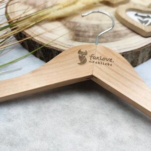 May include: A wooden clothes hanger with a fox logo and the text "foxlove. nochliebe" printed on it. The hanger is on a wooden surface with dried grass.