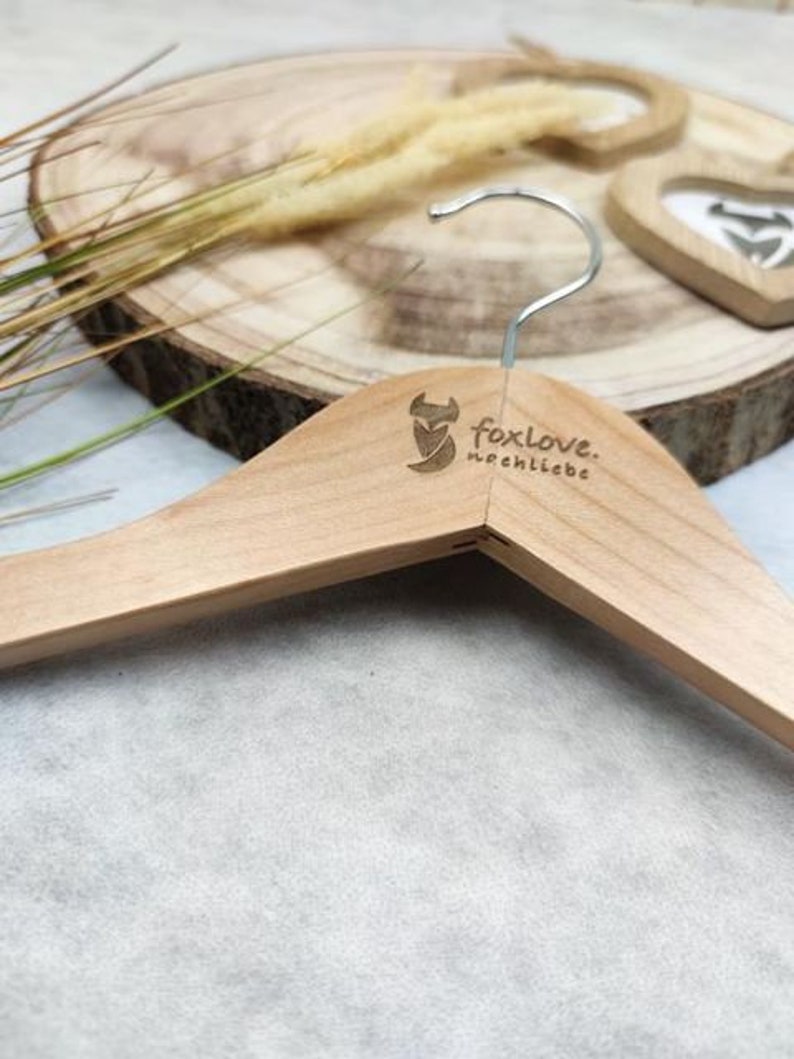 May include: A wooden clothes hanger with a fox logo and the text "foxlove. nochliebe" printed on it. The hanger is on a wooden surface with dried grass.