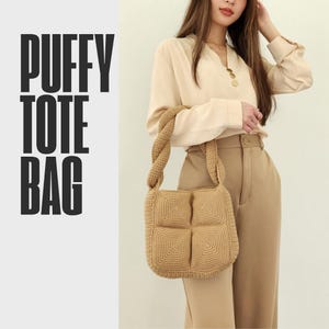 May include: A tan, puffy tote bag with a square, textured design. The bag has a braided handle and the words "PUFFY TOTE BAG" in bold, black letters on the left side. The bag is carried by a person wearing a beige blouse and tan pants.