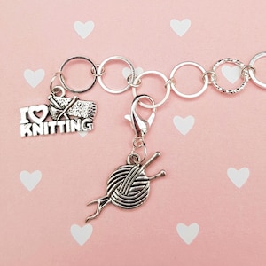 Op de afbeelding: Zilverkleurige bedelarmband met een karabijnsluiting en meerdere bedels. Eén bedel leest "I ❤️ KNITTING" en een andere toont een bol wol met breinaalden. De achtergrond is roze met witte hartvormen.