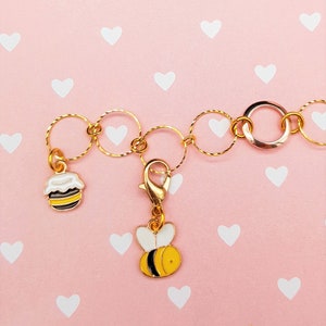 Op de afbeelding: Een gouden ketting met een bij- en een honingpotbedel. De ketting is gemaakt van kleine, met elkaar verbonden cirkels. De bijbedel is geel en zwart, en de honingpotbedel is wit met zwarte strepen.