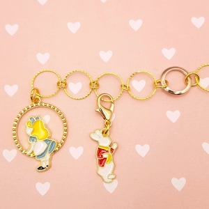 Könnte beinhalten: Ein goldenes Kettenarmband mit zwei Anhängern. Ein Anhänger ist ein Kreis mit einem Cartoon-Bild von Alice aus Alice im Wunderland. Der andere Anhänger ist ein Cartoon-Bild vom weißen Kaninchen.