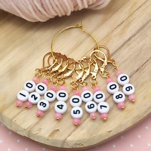 May include: A set of ten gold stitch markers with pink beads and white numbers 0-9. The markers are attached to a gold hoop.