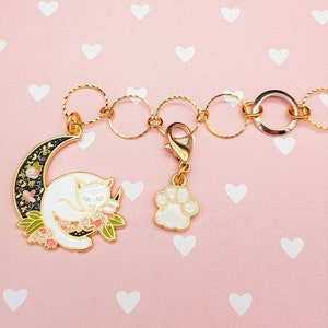Könnte beinhalten: Ein goldfarbenes Armband mit einem weißen Katzen-Anhänger, der auf einem Halbmond-Anhänger schläft, und einem weißen Pfotenabdruck-Anhänger. Die Anhänger sind an einer goldenen Kette mit goldenen Sprungringen befestigt.