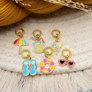 Puede incluir: Una colección de colgantes dorados con diseños de temática veraniega. Los colgantes incluyen un paraguas arcoíris, una bebida tropical, un helado, chanclas, un salvavidas y gafas de sol en forma de corazón. Cada colgante tiene un anillo dorado.