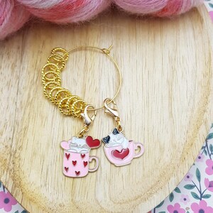 Puede incluir: Dos marcadores de punto dorados con abalorios de esmalte rosa y rojo. Un abalorio es una taza rosa con corazones rojos y un gato, el otro es una taza rosa con un corazón y un gato. Los marcadores de punto están sobre una superficie de madera.