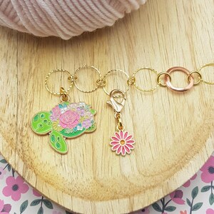 Może przedstawiać: Złoty łańcuszek z wieloma okrągłymi znacznikami ściegów, wisiorkiem w kształcie kwiecistego żółwia i wisiorkiem w kształcie różowego kwiatu. Łańcuszek leży na drewnianej powierzchni.